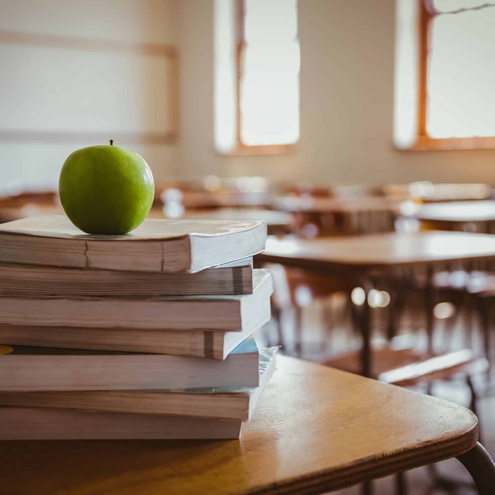Image of a classroom, focusing on a stack of books and an apple on a desk.
